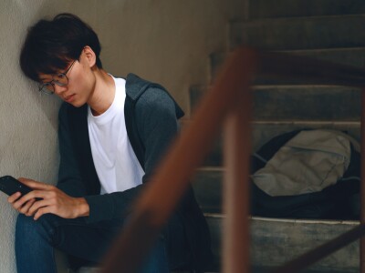 An upset Asian teenage boy student, dressed in casual outfit, sits on a stairway reading on his smartphone with a backpack beside him, while dealing with the issue of cyberbullying.