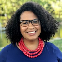 A black woman with shoulder-length curly hair poses for a portrait, smiling directly at the camera. She is wearing glasses and a three-tiered necklace of large red beads on a blue blouse.