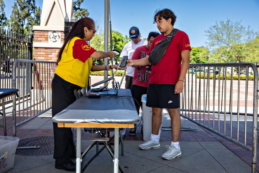 Standing next to some barricades, a woman wearing a red and gold shirt uses a handheld scanning device to scan the phone of a man wearing a red shirt, shorts.