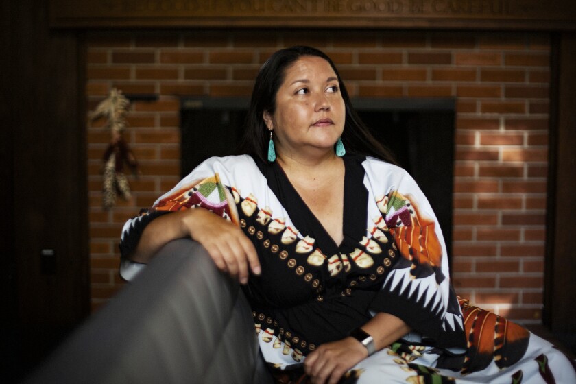 A woman in a bright, colorful gown glances off-camera to the right.  She is sitting on a couch with one  arm resting on the back. She has olive skin, long dark hair, and is wearing large turquoise earrings.