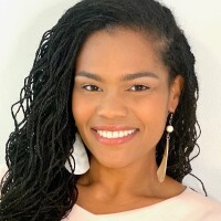 A black woman with long, tight braids smiles for a portrait. She is wearing large earings and a light blouse.