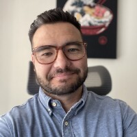 An asian man with a short beard and swept-back hair smiles as he poses for a selfie inside an office. He is wearing large turtle-shell glasses and a grey button-up shirt.
