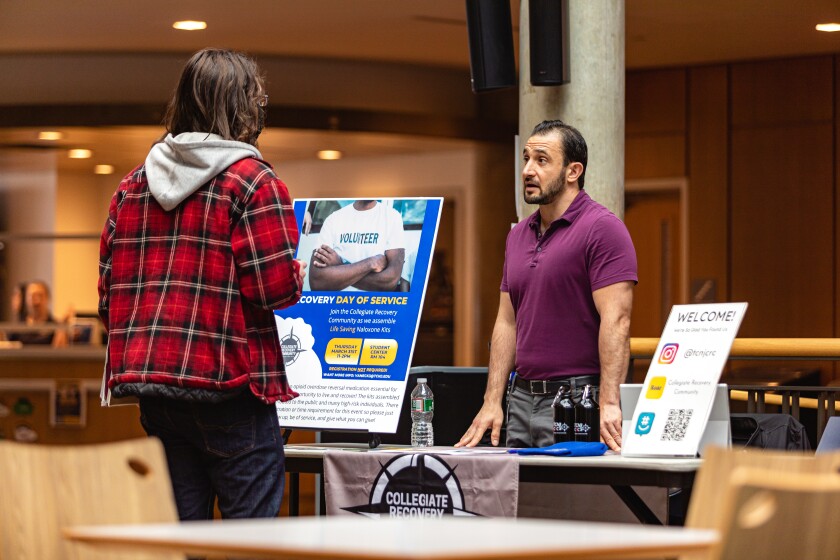 In what looks to be a cafeteria or interior lobby, a bearded white man in casual business attire stands behind an info booth and talks with someone wearing a plaid jacket and jeans with their back to the camera. The booth is a table featuring materials and posters related to drug and alcohol abuse recovery programs.