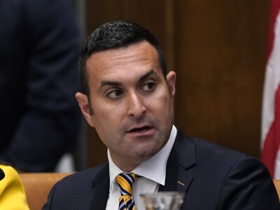 A tan white man is seen in a suit and tie and sporting a short haircut. He looks to the viewer's right. A microphone, infront of him, and a bit of an American flag, behind him, are visible in the image.